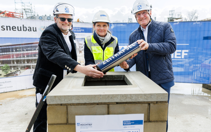 Zwei Männer und eine Frau lassen gemeinsam eine Zeitkapsel in die Baugrube von Activ Sites. Sie tragen Helme und Warnwesten.