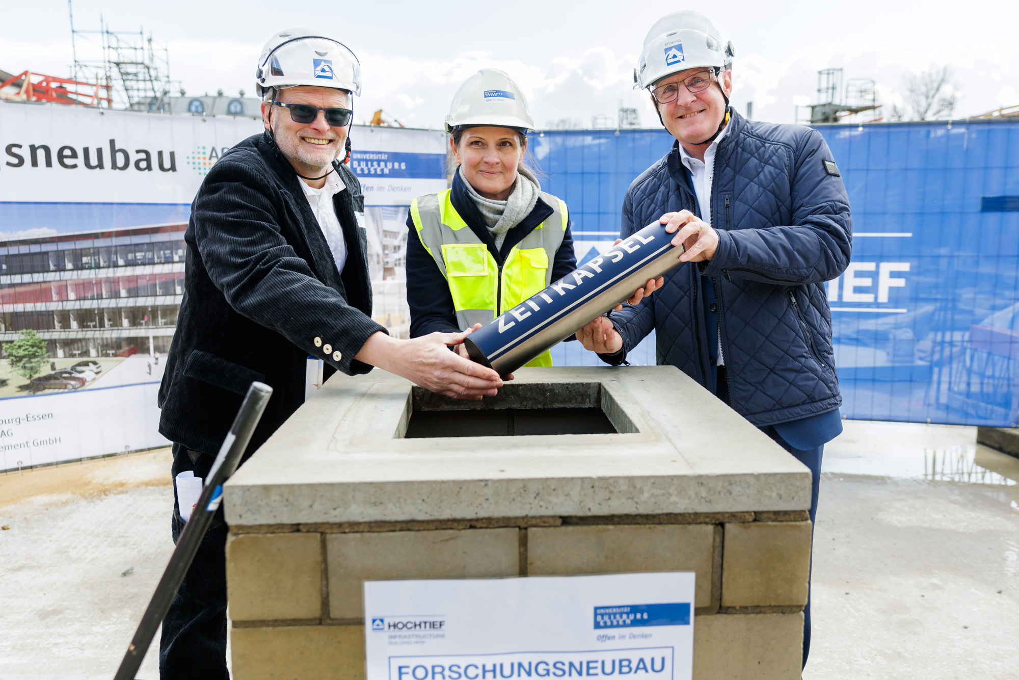 Zwei Männer und eine Frau lassen gemeinsam eine Zeitkapsel in die Baugrube von Activ Sites. Sie tragen Helme und Warnwesten.